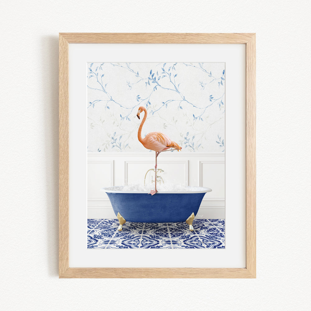 A framed image depicting a pink flamingo standing on the edge of a blue bathtub filled with water, set against a floral wallpaper background.