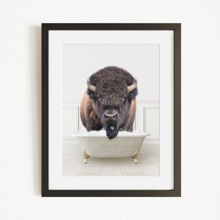 A buffalo with a black mane and horns is sitting in a white bathtub, with its head in the water.
