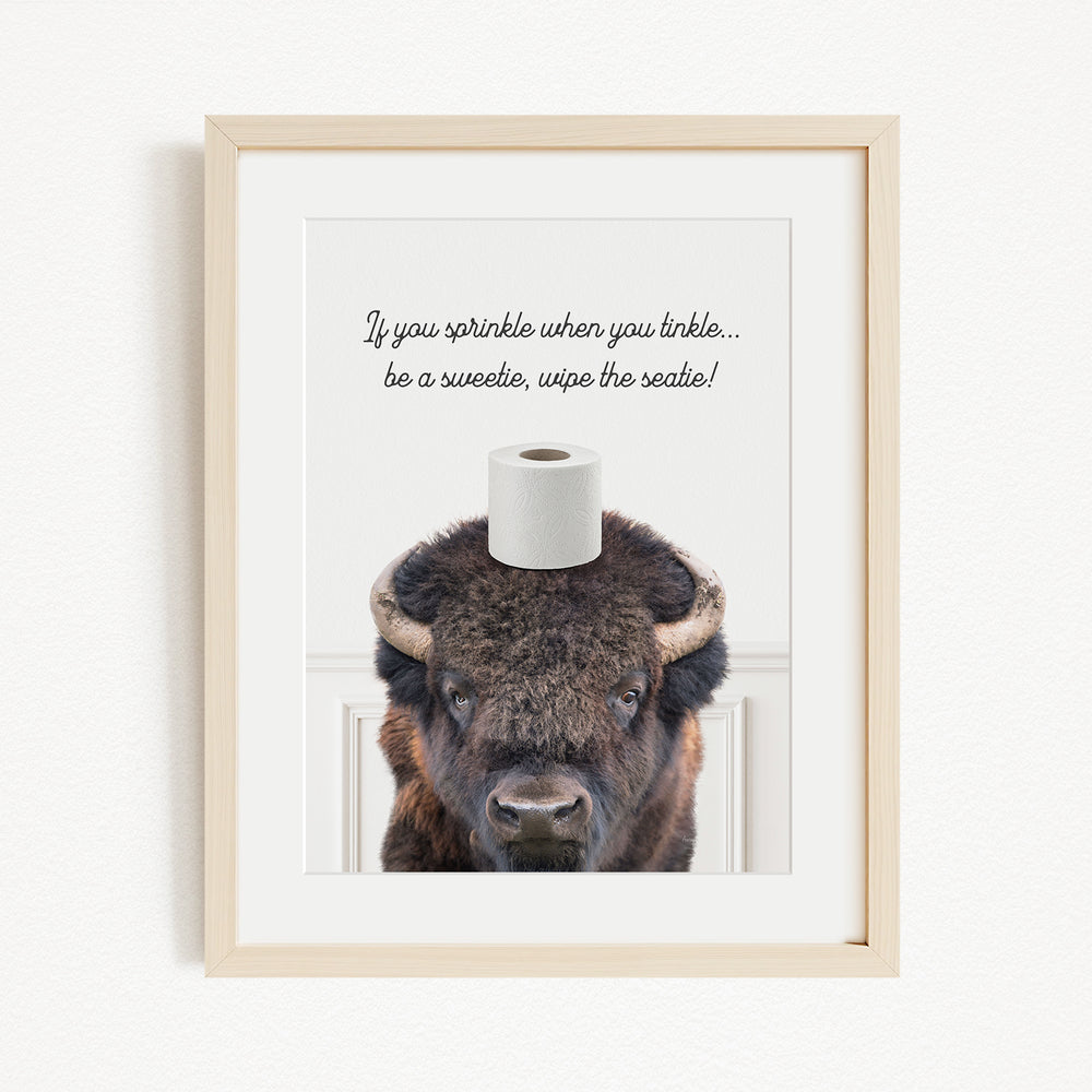 A framed image of a bison with a roll of toilet paper on its head, accompanied by a humorous text that reads "If you sprinkle when you linkle... be a sweetie, wipe the seat!"