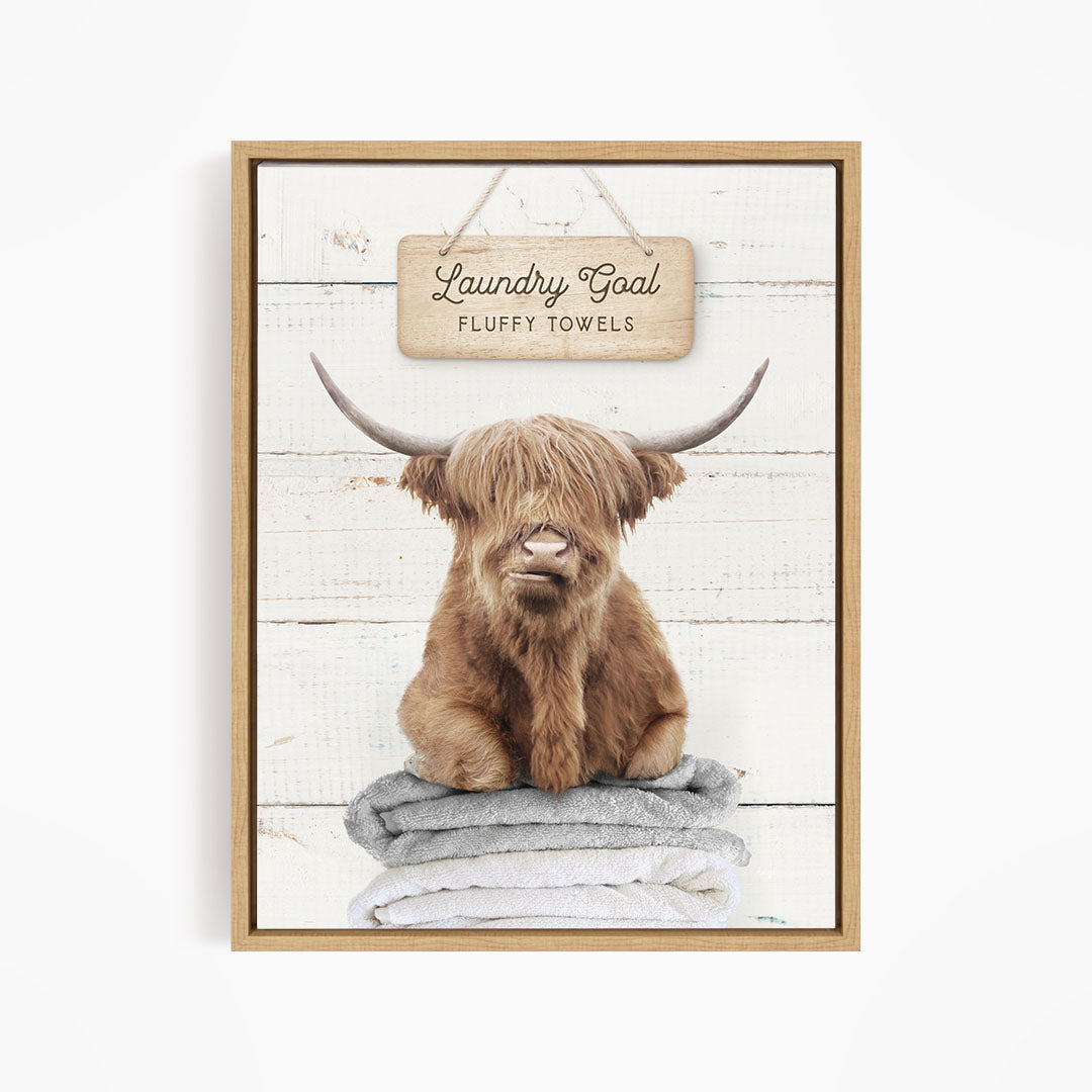 A framed image of a brown cow with long horns sitting on a stack of fluffy towels, with a sign above that says "Laundry Goal Fluffy Towels".