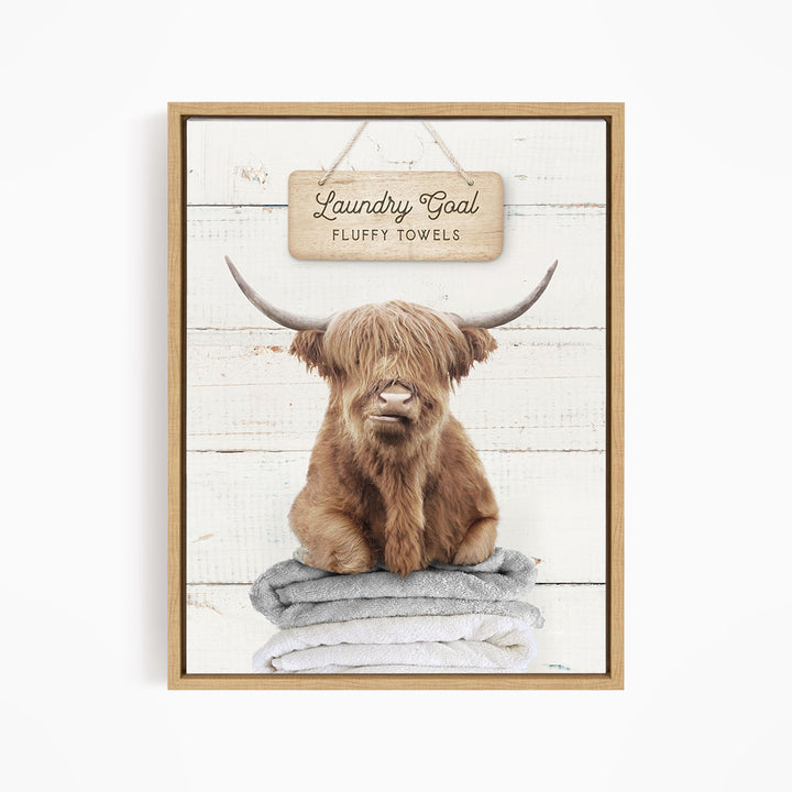 A framed image of a brown cow with long horns sitting on a stack of fluffy towels, with a sign above that says "Laundry Goal Fluffy Towels".