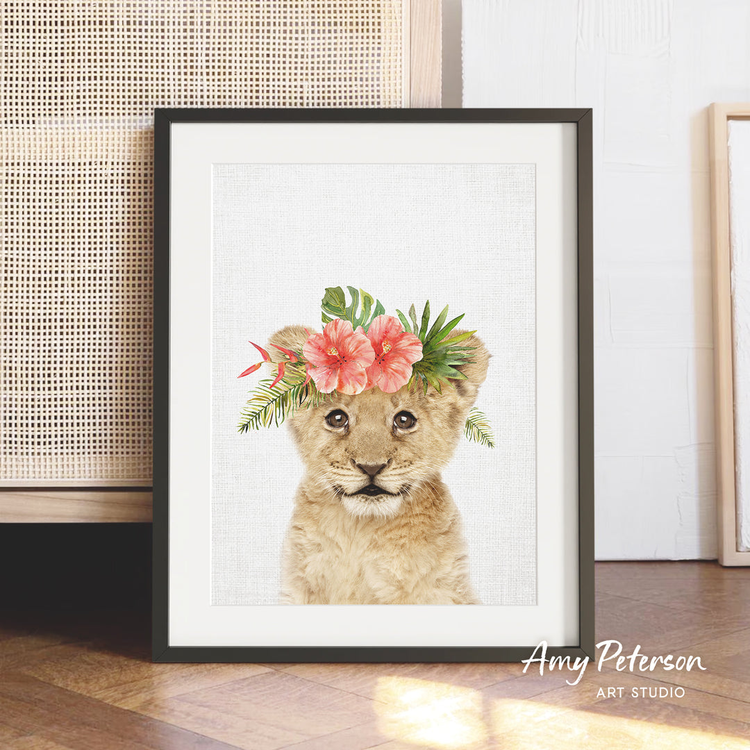 A framed print of a young lion cub wearing a floral headpiece is displayed on a wooden floor against a white wall.