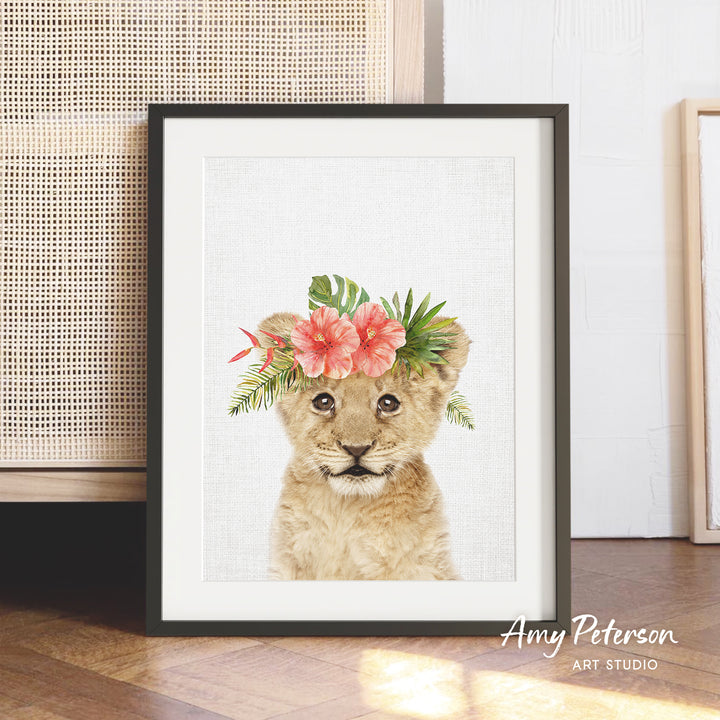 A framed print of a young lion cub wearing a floral headpiece is displayed on a wooden floor against a white wall.