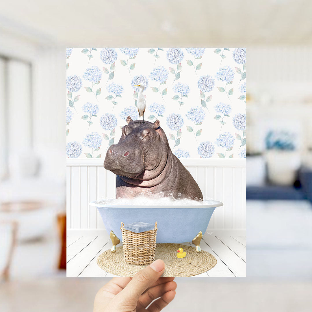 A hand holds up a framed image depicting a hippopotamus sitting in a bathtub filled with bubbles, with a bird perched on its head.