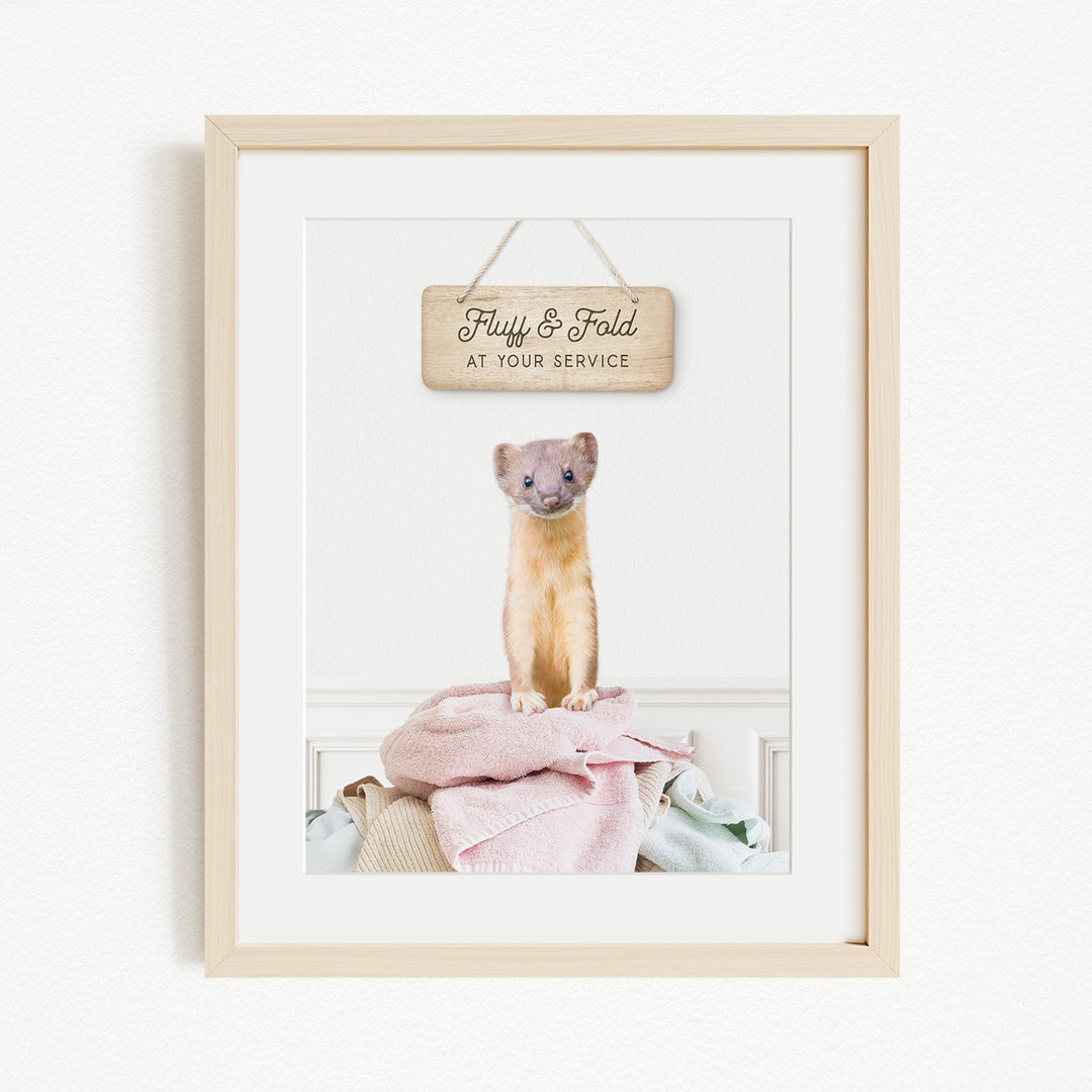 A framed image of a small cat sitting on a pile of clothes, with a wooden sign above it that says "Fluffy & Fold at your service".