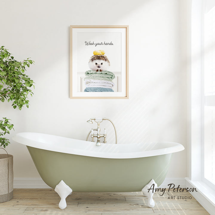 A framed poster with a hedgehog sitting on a stack of towels hangs on a white wall above a green clawfoot bathtub.