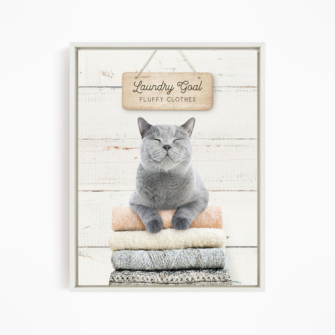 A gray cat sitting on a stack of folded clothes, with a wooden sign above that says "Laundry Goal Fluffy Clothes".