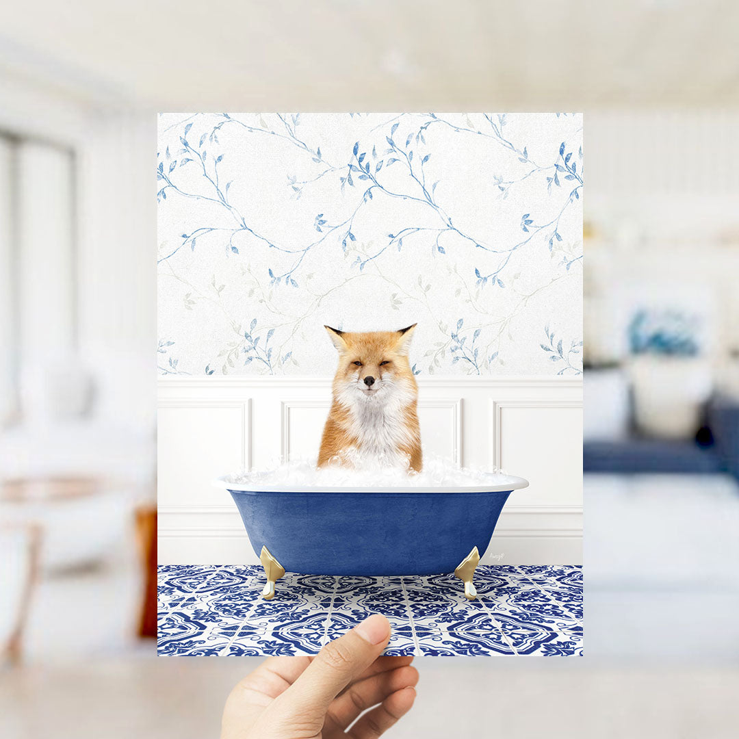 A hand holds up a framed image of a fox sitting in a blue bathtub filled with bubbles, set against a floral wallpaper backdrop.