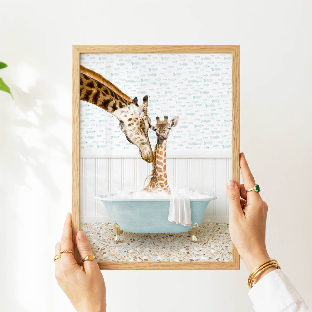 A person's hands are holding a framed picture of two giraffes in a bathtub, with one giraffe appearing to be kissing the other giraffe.