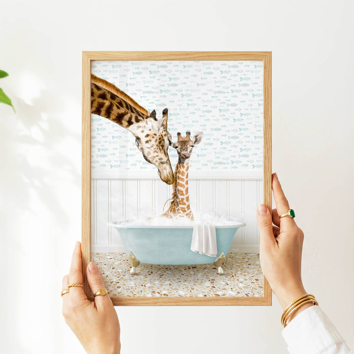 A person's hands are holding a framed picture of two giraffes in a bathtub, with one giraffe appearing to be kissing the other giraffe.