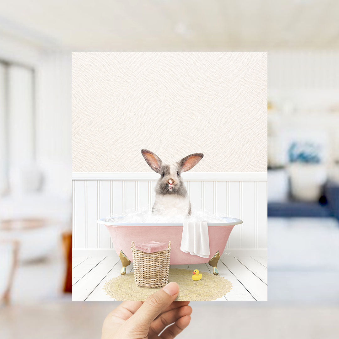 A hand holds up a framed image depicting a rabbit sitting in a pink bathtub filled with bubbles, surrounded by a yellow rubber duck and a white towel.