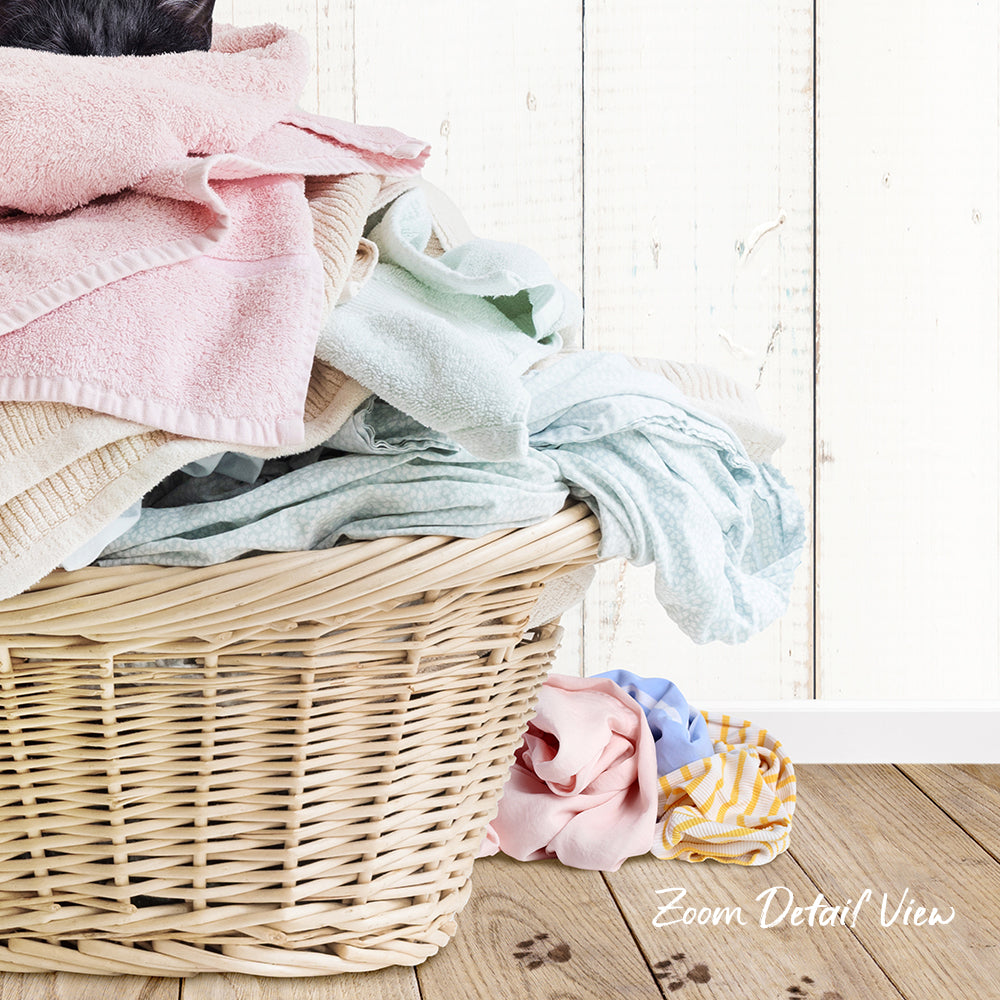 A wicker basket filled with various laundry items, including towels and clothes, sits on a wooden floor against a white wooden wall.