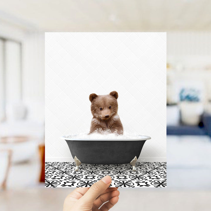 A hand holds up a framed image of a small brown bear sitting in a bathtub filled with bubbles, creating a whimsical and playful scene.