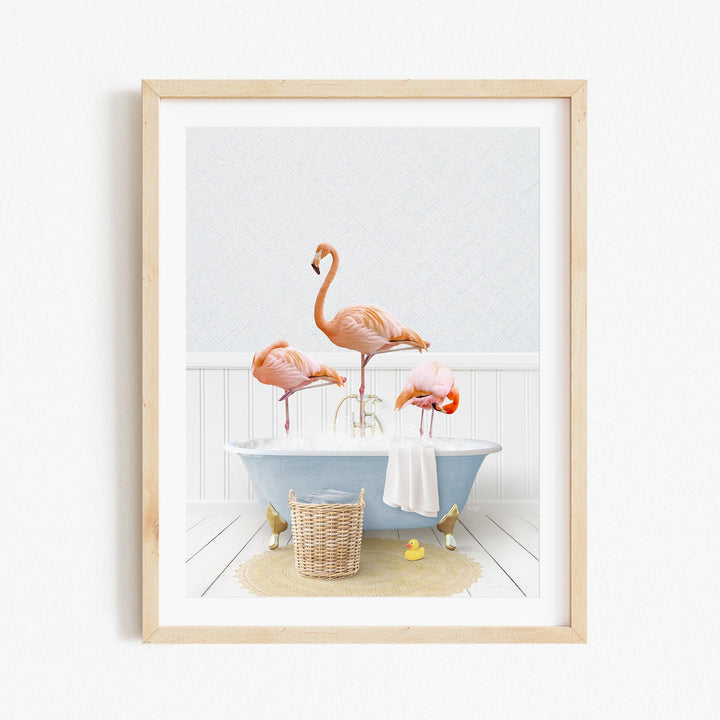 A framed image depicting three flamingos standing in a bathtub filled with water, surrounded by a basket of bath toys and a towel.