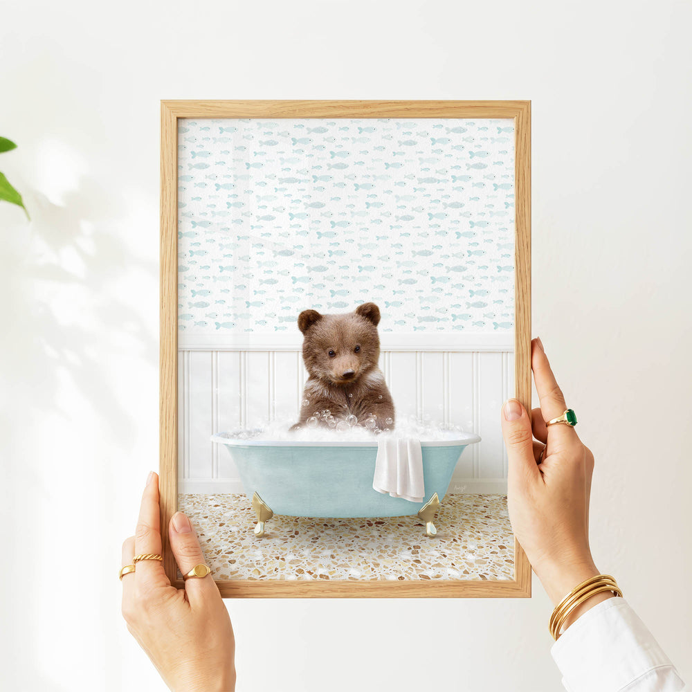 A person's hands are holding a framed picture of a small brown bear sitting in a blue bathtub filled with water, surrounded by a pattern of fish.