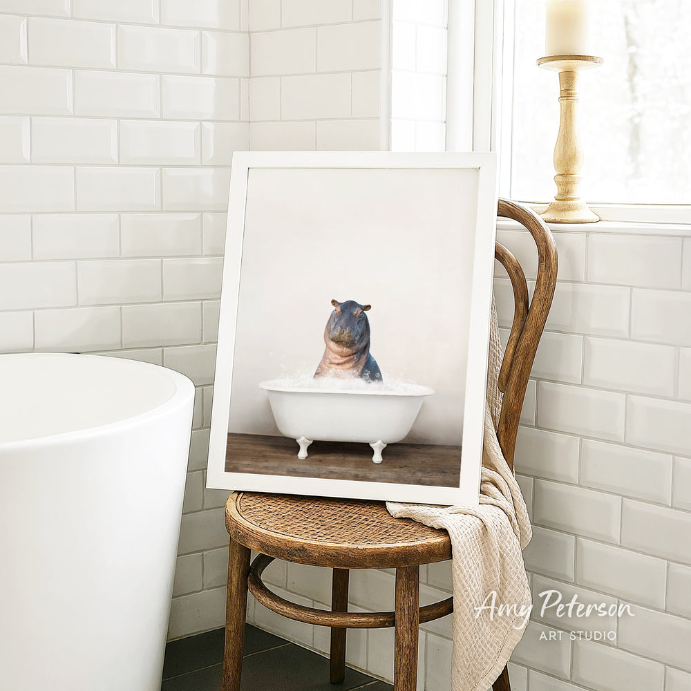 A framed image of a hippopotamus sitting in a bathtub is displayed on a wooden chair in a bathroom setting.