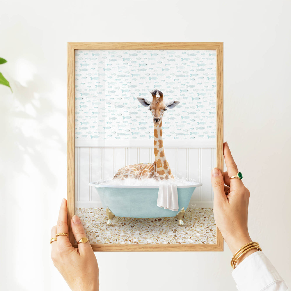 A person's hands are holding a framed picture of a giraffe sitting in a blue bathtub filled with water, surrounded by fish-themed wallpaper.