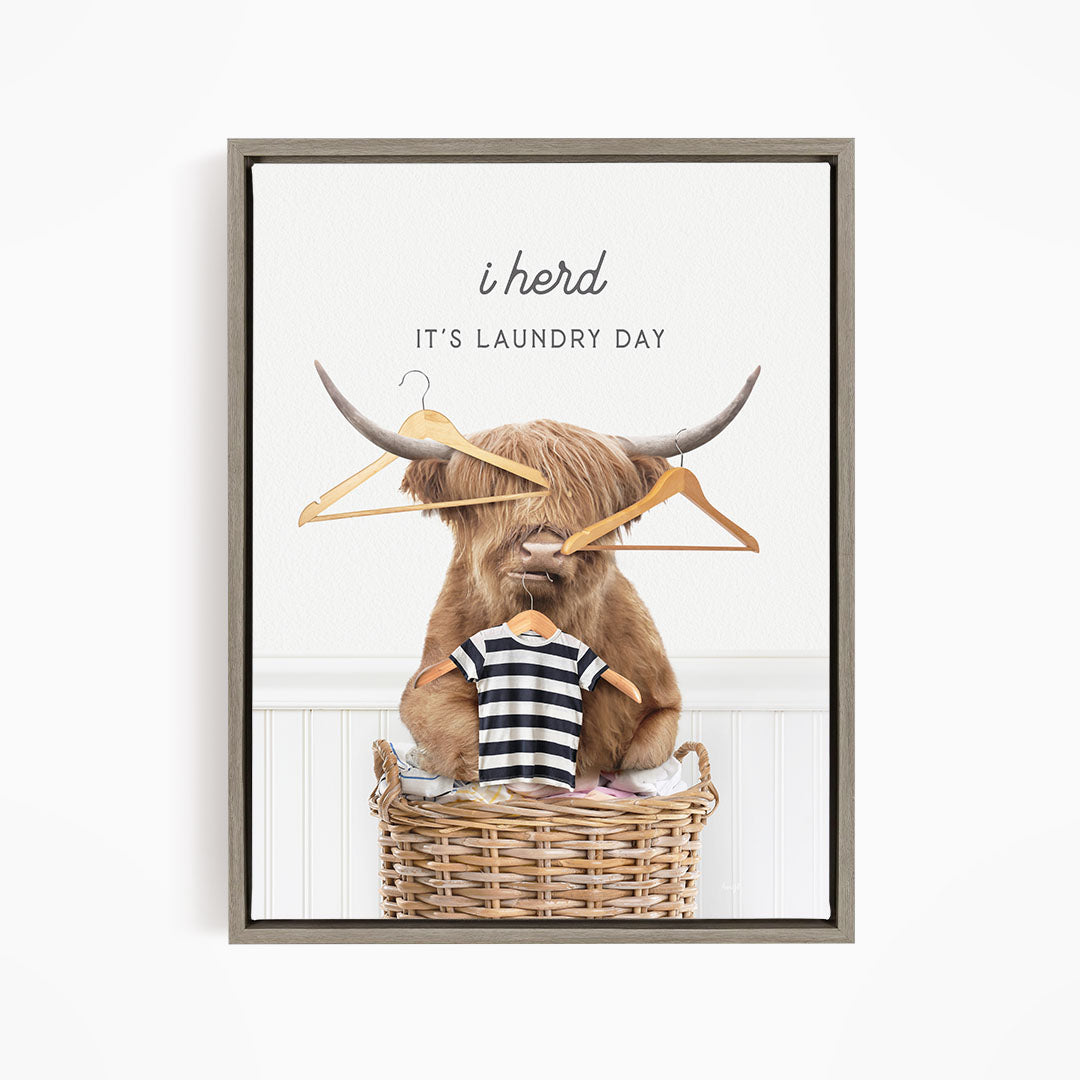 A framed image depicting a Highland cow sitting in a laundry basket, with clothes hanging from the baskets and a sign above the cow that reads "It's laundry day".