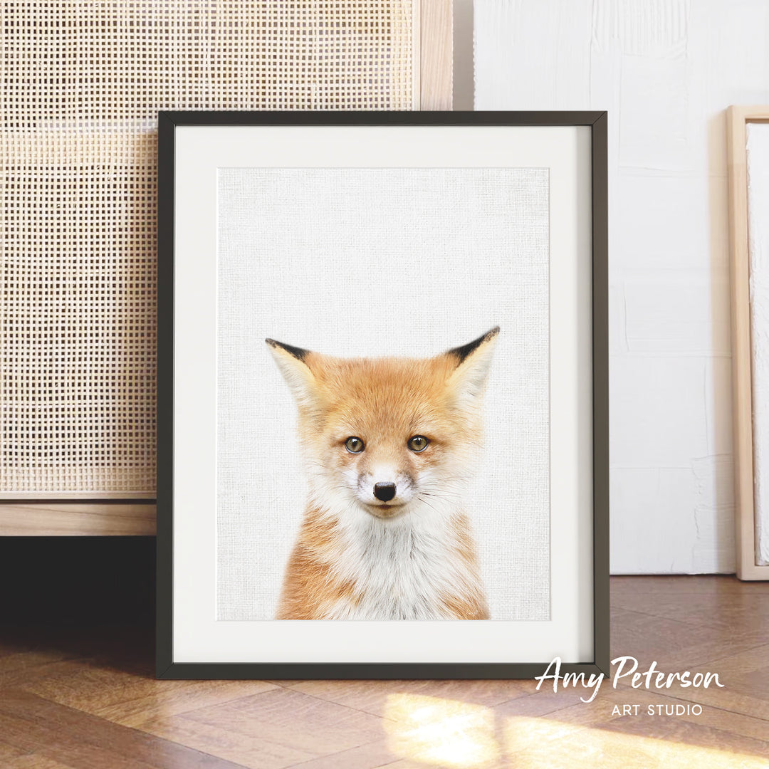 A framed print of a young fox with a white background and a black border, displayed on a wooden floor against a white wall.