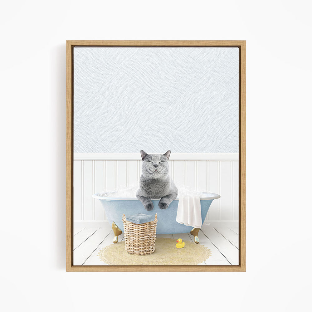 A gray cat sitting in a blue bathtub filled with bubbles, surrounded by a yellow rubber duck and a wicker basket.