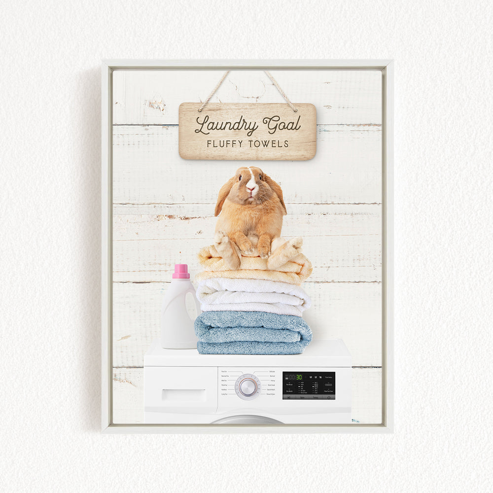A wooden sign with the text "Laundry Goal Fluffy Towels" hangs above a stack of folded towels and a washing machine.