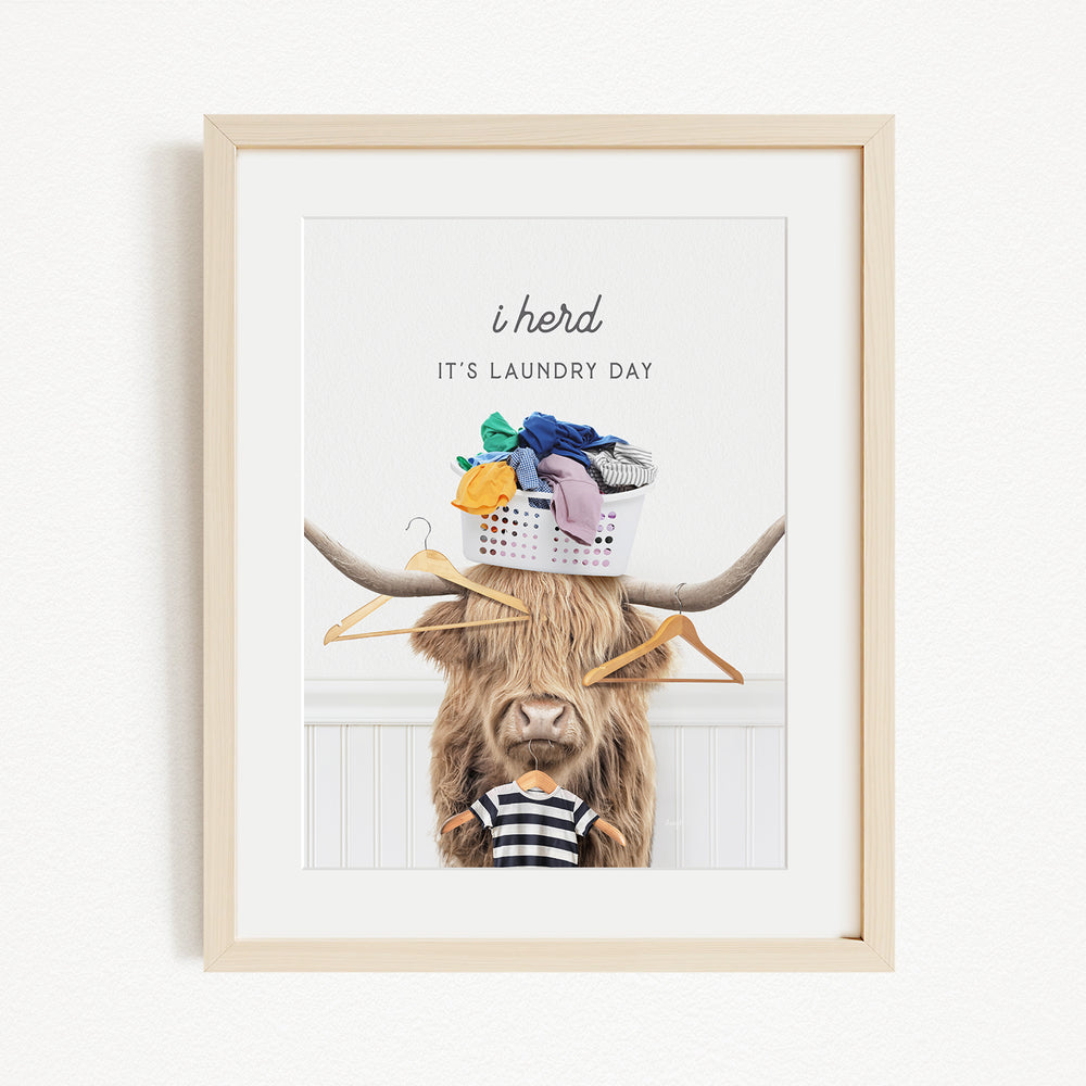 A framed image depicting a cow with a basket of laundry on its head, accompanied by the text "It's laundry day" and a playful caption "It's laundry day" with a cow wearing a shirt.