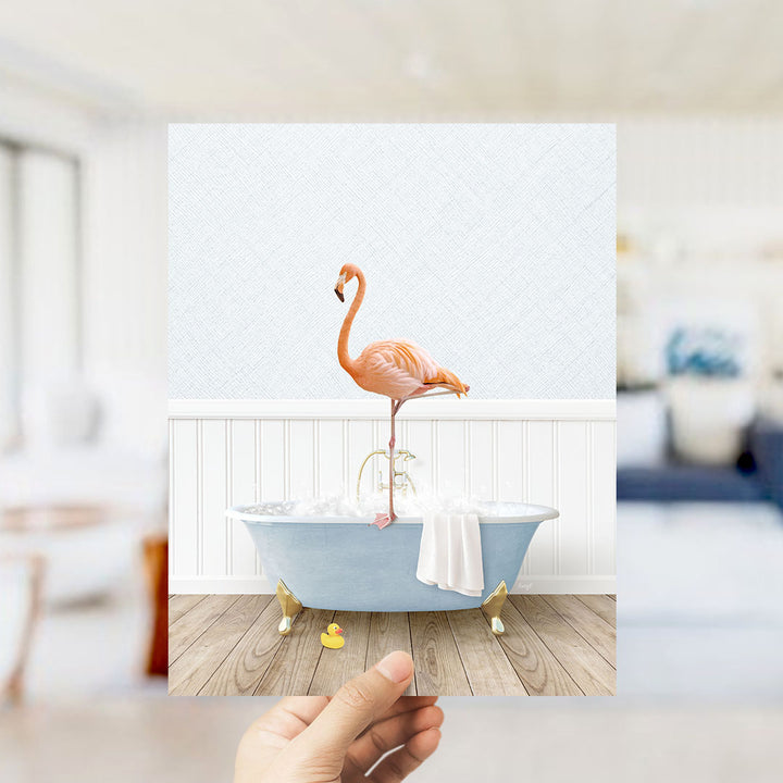 A hand holding a photograph of a pink flamingo standing on a blue bathtub filled with bubbles, with a white wall and wooden floor in the background.