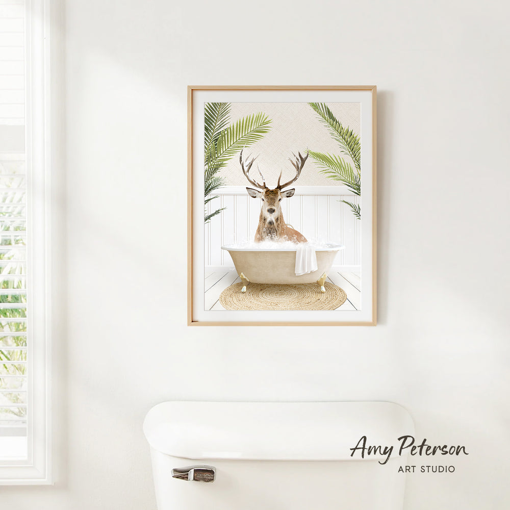 A framed image of a deer sitting in a bathtub, surrounded by palm leaves, is hanging on a white wall in a bathroom.