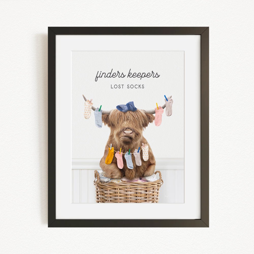 A framed image depicting a brown cow sitting in a wicker basket filled with colorful socks hanging from clotheslines.