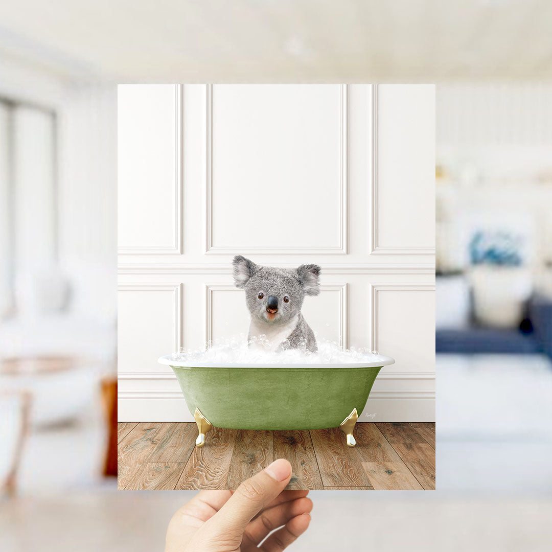 A hand holds up a photograph of a koala bear sitting in a green bathtub filled with bubbles, creating a whimsical and playful scene.