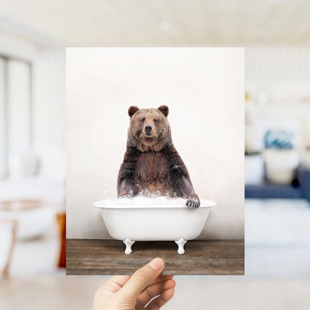 A hand holds up a framed image of a brown bear sitting in a white bathtub filled with water, creating a whimsical and playful scene.