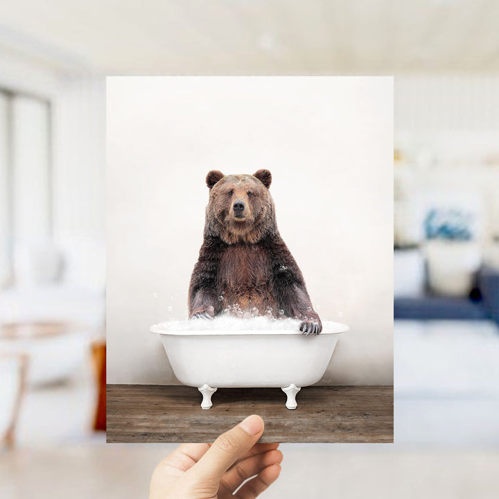 A hand holds up a framed image of a brown bear sitting in a white bathtub filled with water, creating a whimsical and playful scene.