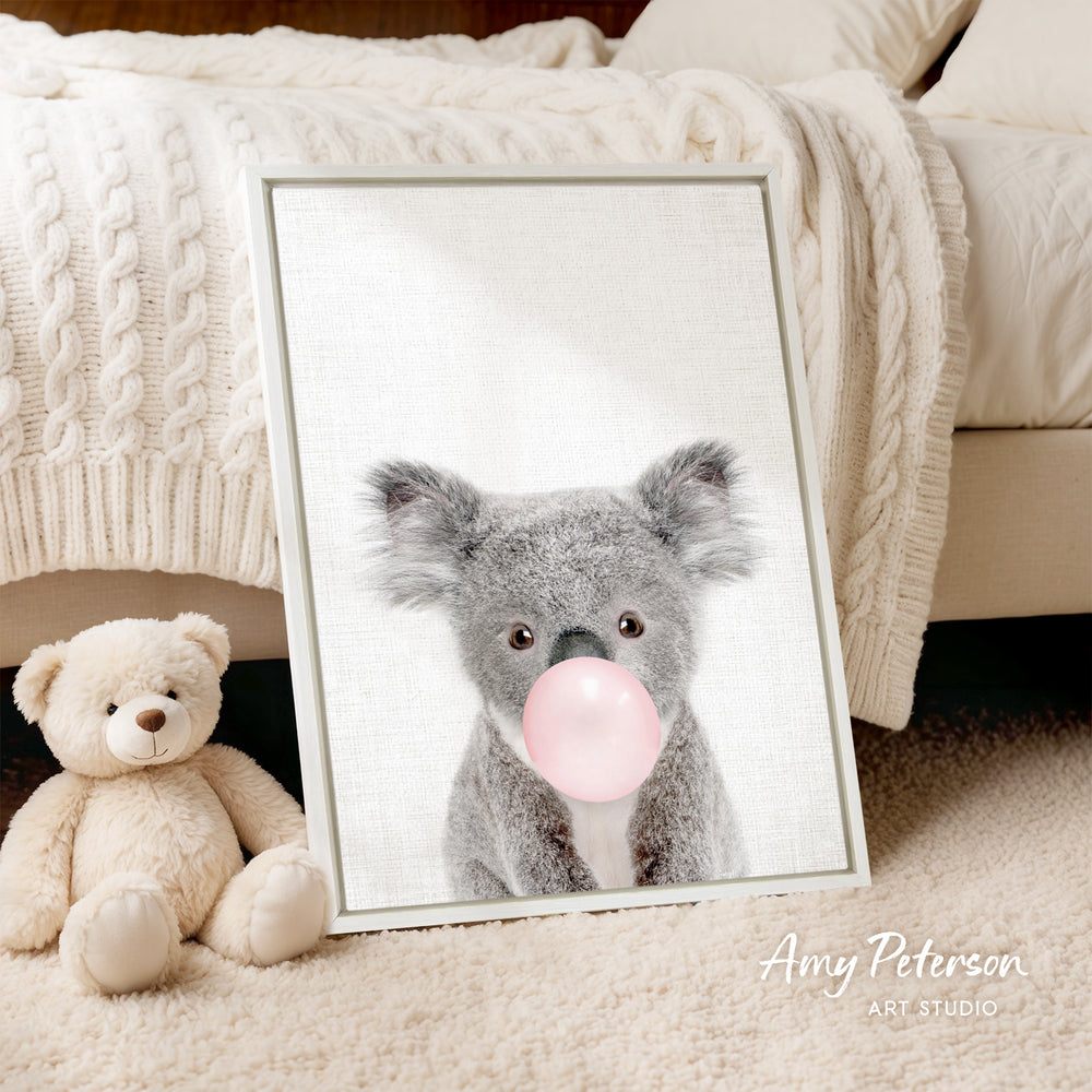 A framed image of a koala bear blowing a bubble gum bubble is displayed on a bed, accompanied by a teddy bear.