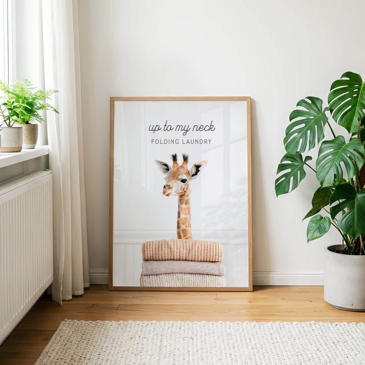 a framed poster of a giraffe with the text "up to my neck" and "Folding laundry" on it, placed on a wooden floor next to a potted plant.