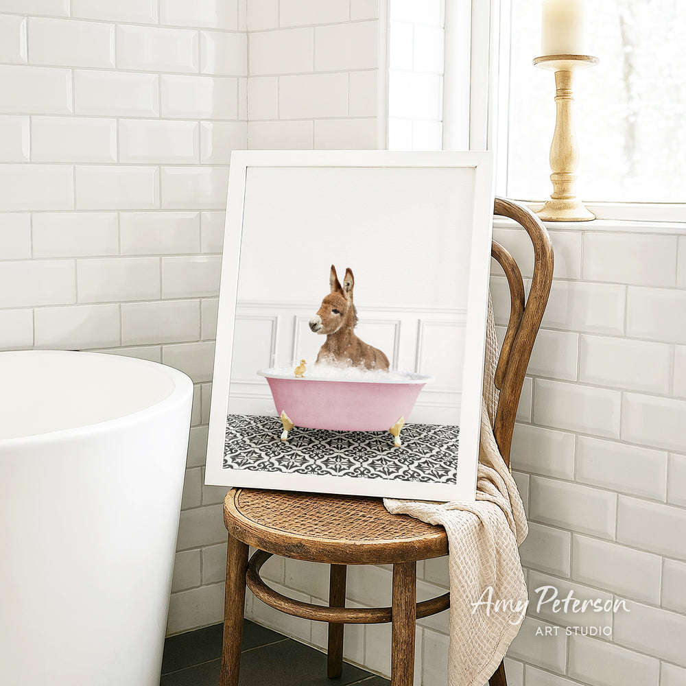 A framed image of a small dog sitting in a pink bathtub, with a towel draped over the back of a wooden chair in a bathroom setting.
