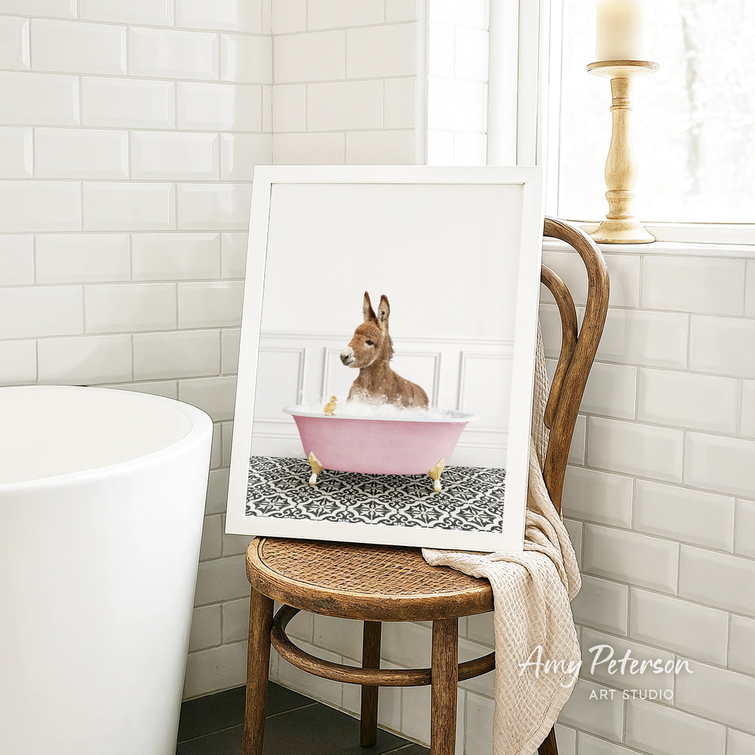 A framed image of a small dog sitting in a pink bathtub, with a towel draped over the back of a wooden chair in a bathroom setting.