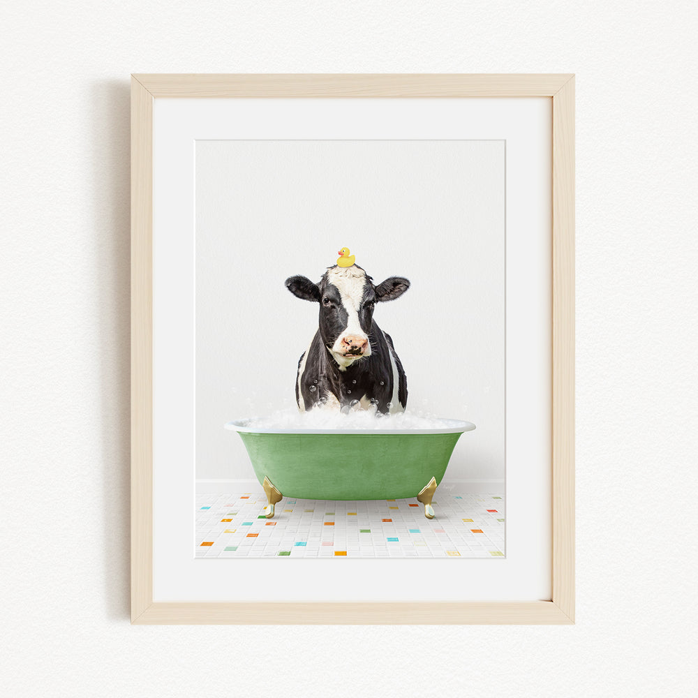 A black and white cow is sitting in a green bathtub filled with bubbles, with a yellow duck perched on its head.