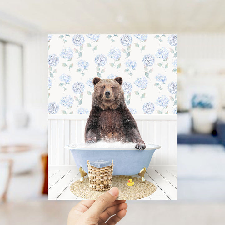 A hand holds up a framed image of a brown bear sitting in a blue bathtub filled with bubbles, surrounded by a floral patterned wall.