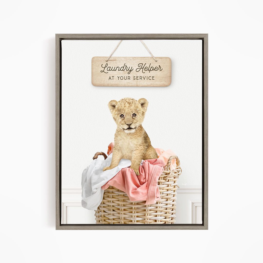 A young lion cub sitting in a wicker basket filled with clothes, with a wooden sign above that reads "Laundry Helper at your service".