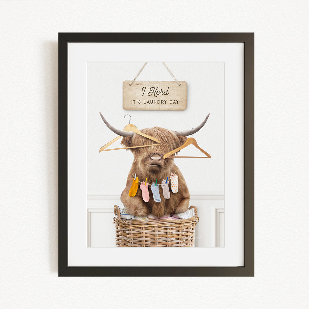 A framed image depicting a cow with long horns sitting in a wicker basket, surrounded by clothes and a sign that reads "I herd it's laundry day".