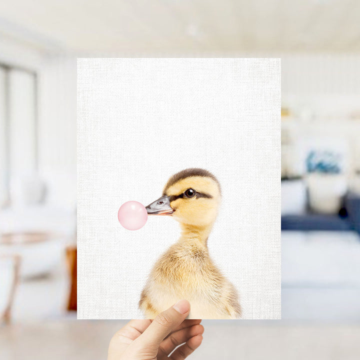A person's hand is holding up a white square with a photograph of a duck blowing a bubble with a pink bubble gum bubble.