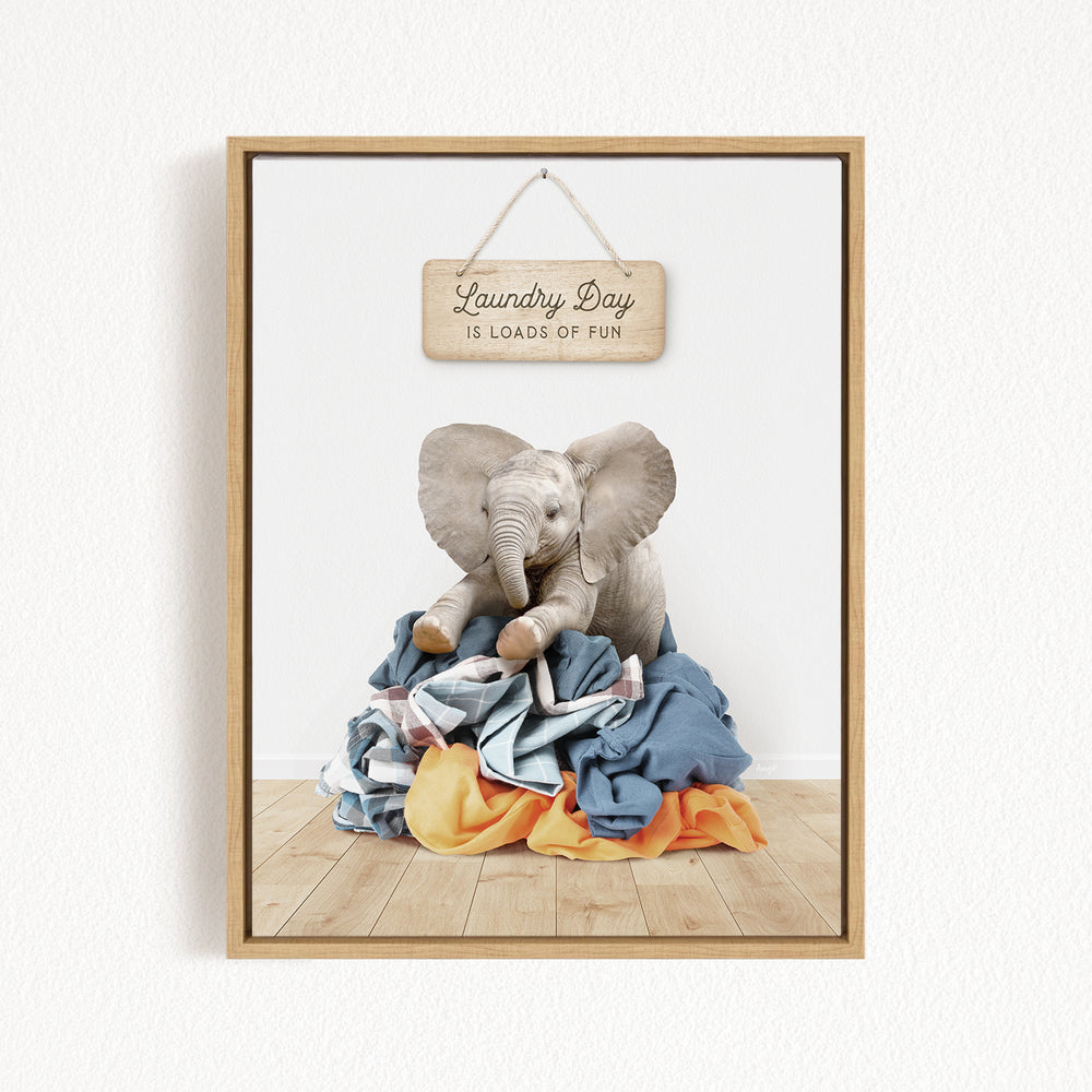 A framed image of a stuffed elephant sitting atop a pile of laundry, with a wooden sign above that reads "Laundry Day is loads of fun".