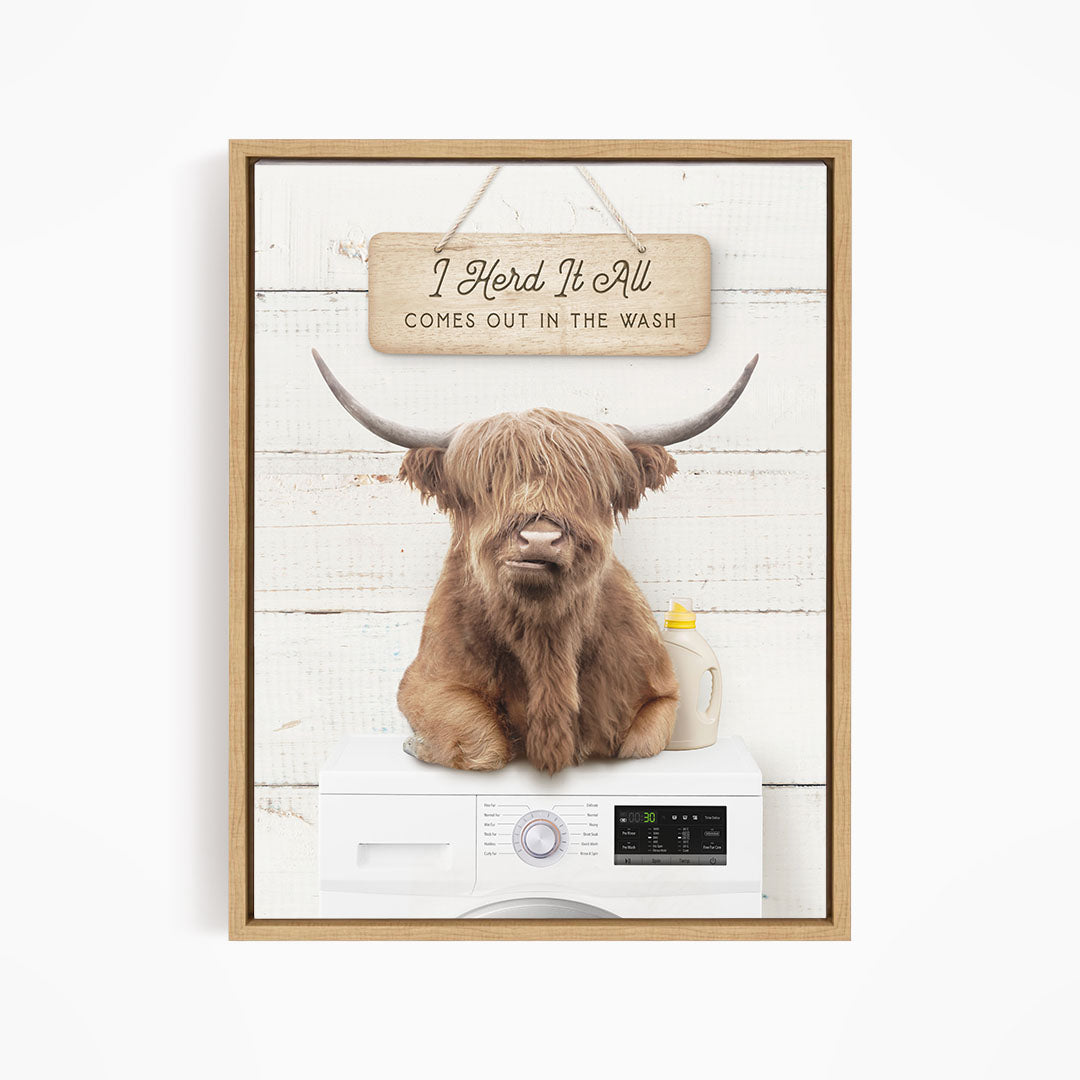 A framed image depicting a large, shaggy-haired animal, possibly a yak or a bison, sitting on top of a washing machine. The animal appears to be looking directly at the viewer, with a sign above it that reads "I Herd It All Comes out in the Wash".