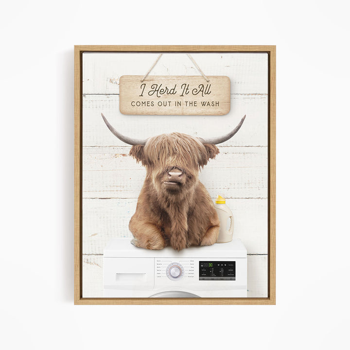A framed image depicting a large, shaggy-haired animal, possibly a yak or a bison, sitting on top of a washing machine. The animal appears to be looking directly at the viewer, with a sign above it that reads "I Herd It All Comes out in the Wash".