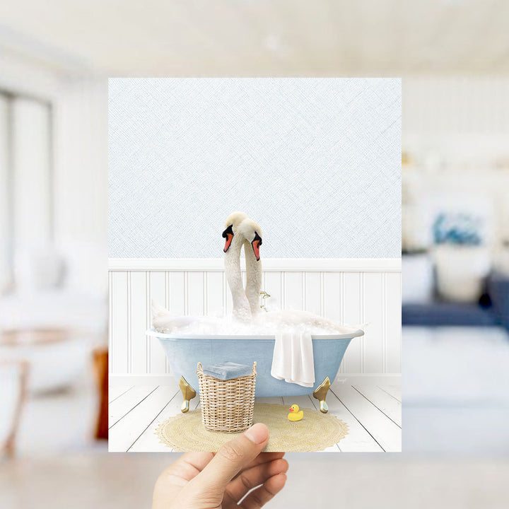 A hand holds up a framed image depicting two swans bathing in a blue bathtub, with a basket of rubber ducks nearby.