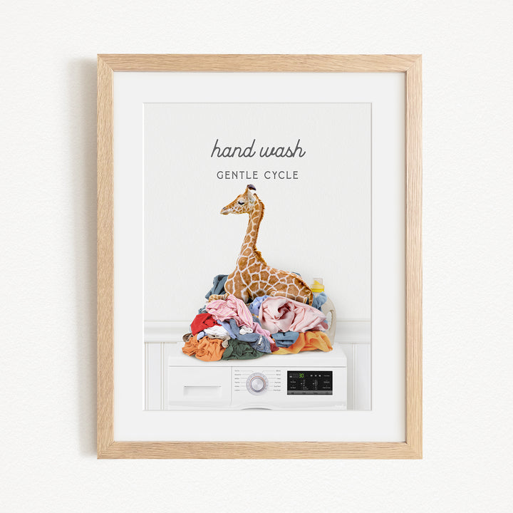 A framed image depicting a giraffe sitting atop a pile of laundry, with the text "hand wash gentle cycle" written above it.