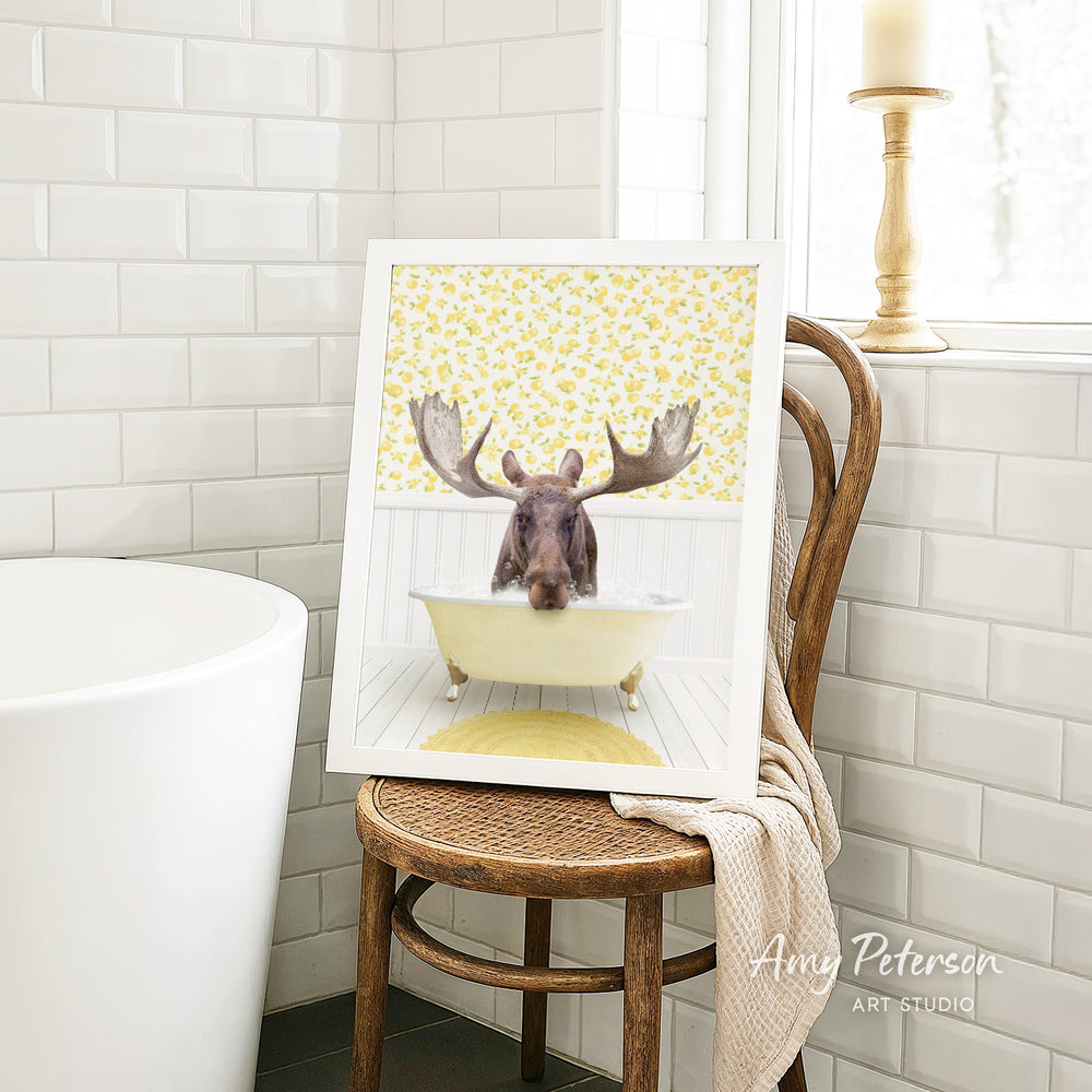 A framed image of a moose sitting in a bathtub, with its antlers raised, is displayed on a wooden chair in a bathroom setting.