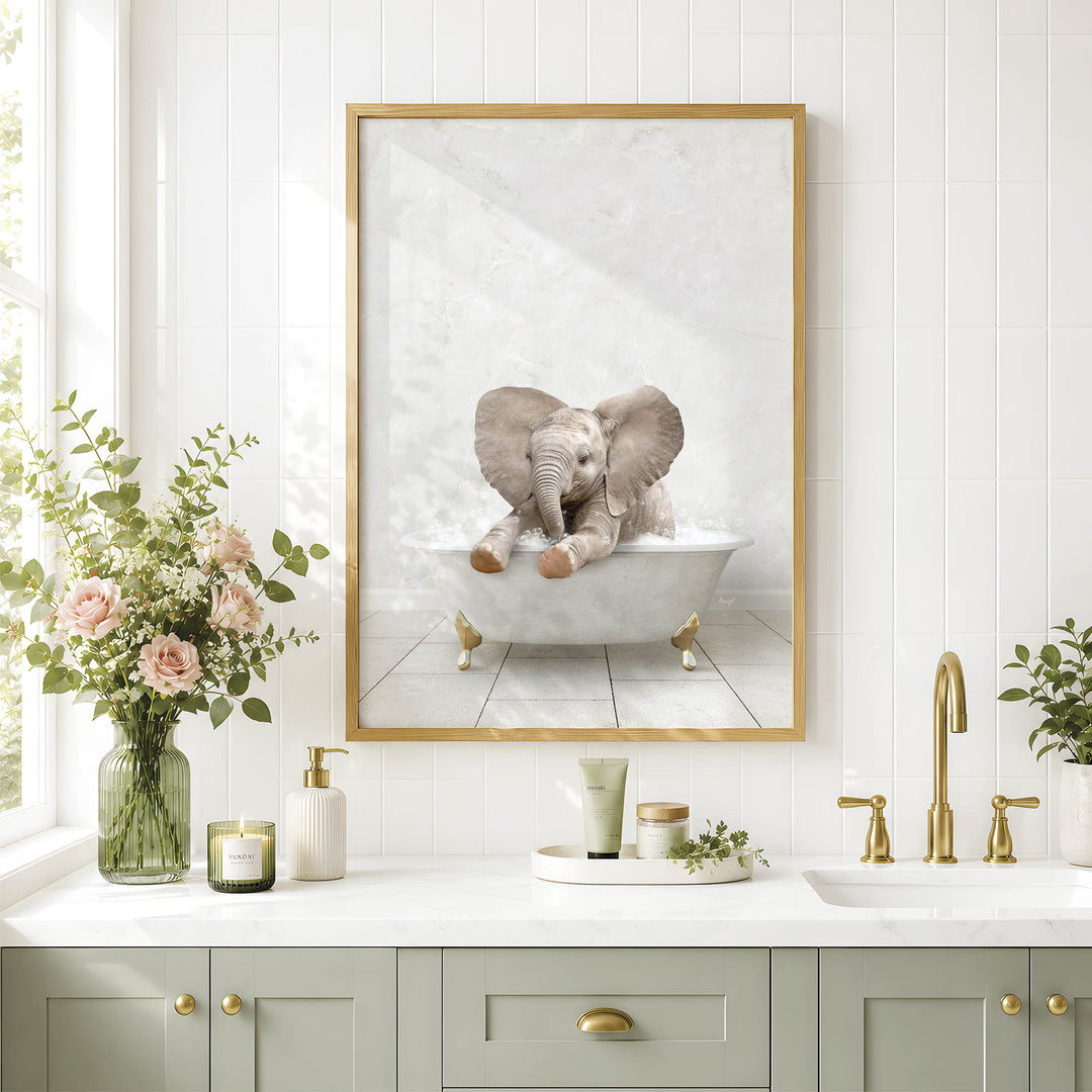 A framed image of a baby elephant sitting in a bathtub is displayed on the wall above a bathroom counter with various toiletries and a vase of flowers.