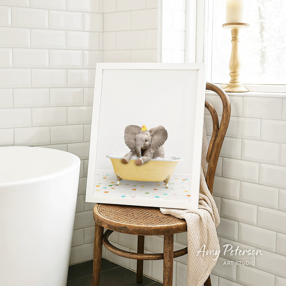 A framed image of a baby elephant sitting in a bathtub, surrounded by colorful tiles, is displayed on a wooden chair in a bathroom setting.