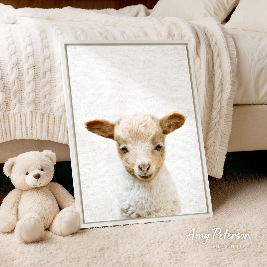 A framed image of a lamb peeking out from behind a white blanket, accompanied by a teddy bear on a beige carpet.